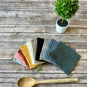 May include: A selection of colourful, crocheted square potholders in various shades, including pink, yellow, and blue. A wooden spoon and a small potted plant are also visible on a wooden surface.