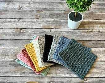 May include: A selection of colourful, crocheted square potholders in various shades, including pink, yellow, and blue. A wooden spoon and a small potted plant are also visible on a wooden surface.