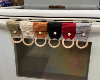 May include: A row of seven handmade crochet kitchen towel holders in various colors, including olive green, cream, orange, black, red, and gray. Each holder has a wooden ring and a wooden button with the word "hope" or "love".