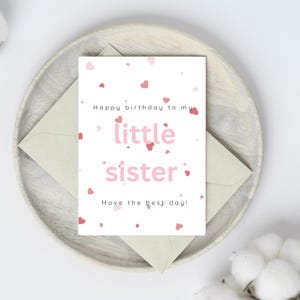 May include: A birthday card with the message "Happy birthday to my little sister" in pink lettering, surrounded by small red hearts. The card is on a wooden plate with a light green envelope.