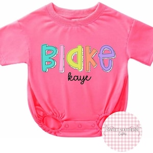 May include: A bright pink short-sleeved romper featuring the name Blake in large, colorful, outlined bubble letters with the name kaye written underneath in a smaller black script font. The garment has a round neckline and snap closures.