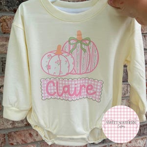 May include: A pale yellow long-sleeve romper featuring a graphic of two pumpkins with pink floral and striped patterns, accented by a green bow. Below the pumpkins, a pink and white gingham scalloped rectangle displays the name Claire in pink.