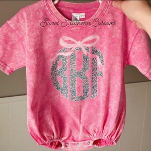 May include: A pink short-sleeved baby romper with a floral monogram and bow design. The romper has the text "Sweet Southern Customs" printed on the top. The romper is size 6-12 months.