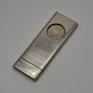 May include: A silver metal matchbox with a circular design on the top. The design features a compass with the letters N, E, S, and W.