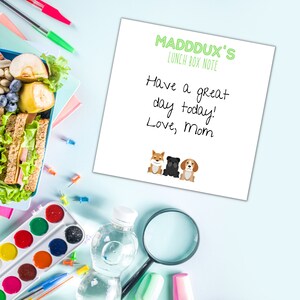 May include: A white lunchbox note with green text that reads "Madddux's Lunch Box Note" and "Have a great day today! Love, Mom". Three cartoon dogs are at the bottom of the note. The dogs are a brown and white Shiba Inu, a black pug, and a brown and white beagle.