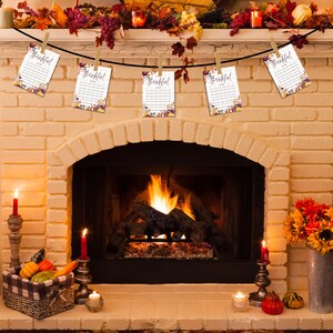 Puede incluir: Una repisa de chimenea decorada con una guirnalda de hojas de oto&ntilde;o y bayas. Cinco tarjetas blancas con el texto "Agradecido Por" est&aacute;n colgadas a trav&eacute;s de la repisa con pinzas para la ropa. La chimenea est&aacute; encendida con fuego.