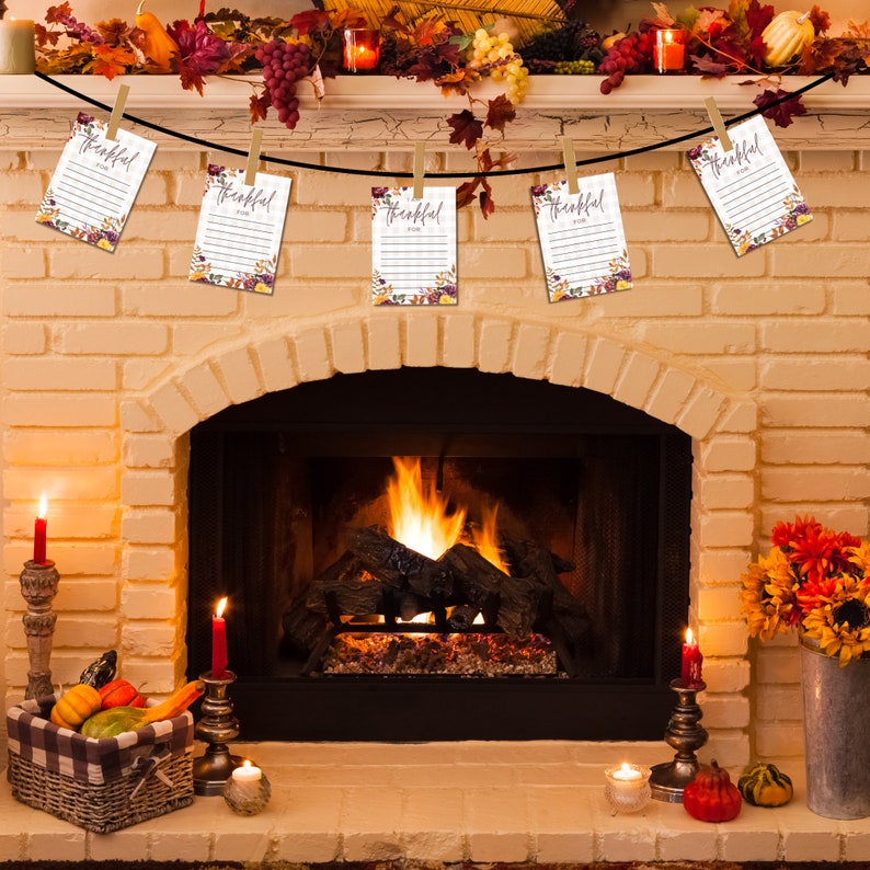 Puede incluir: Una repisa de chimenea decorada con una guirnalda de hojas de oto&ntilde;o y bayas. Cinco tarjetas blancas con el texto "Agradecido Por" est&aacute;n colgadas a trav&eacute;s de la repisa con pinzas para la ropa. La chimenea est&aacute; encendida con fuego.