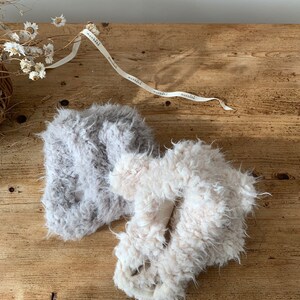 May include: Two fluffy, textured slippers in light gray and off-white, resting on a rustic wooden surface. A ribbon with the word "monbēbē" is draped across the top. Dried flowers add a delicate touch to the scene.