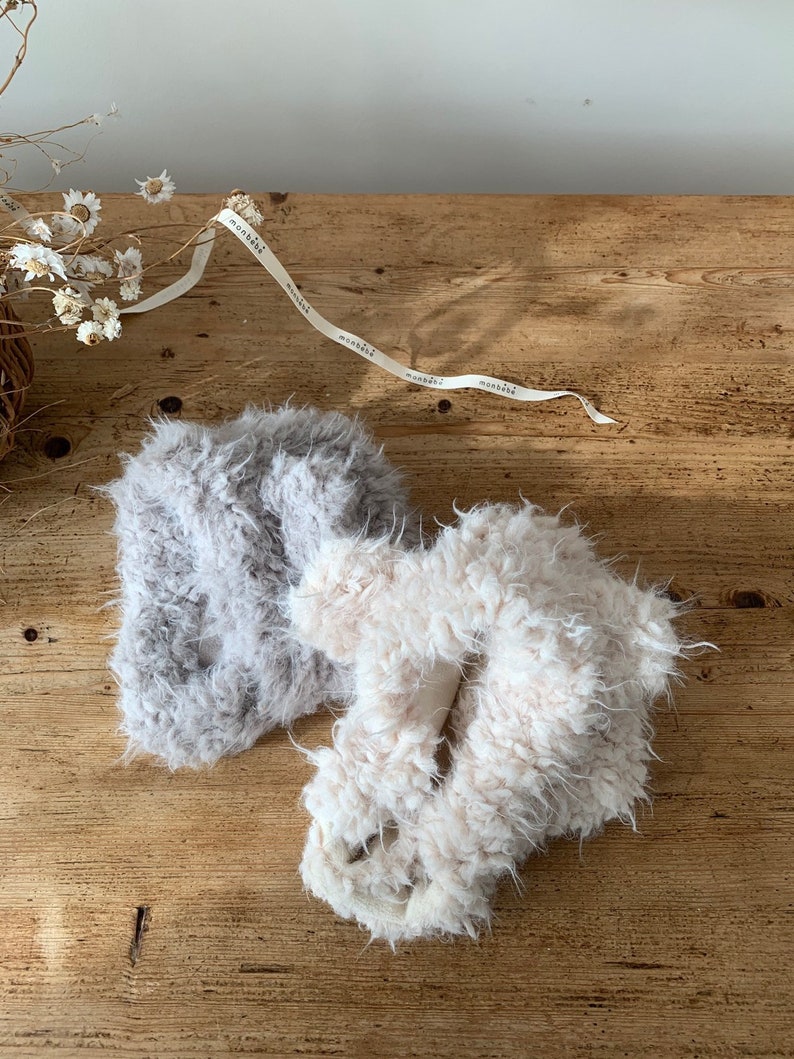 May include: Two fluffy, textured slippers in light gray and off-white, resting on a rustic wooden surface. A ribbon with the word "monbēbē" is draped across the top. Dried flowers add a delicate touch to the scene.