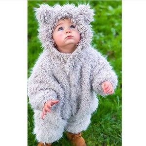 May include: A baby wearing a gray, furry bear costume with a hood and brown boots stands on green grass. The costume has a button closure and bear ears on the hood. The baby is looking up.
