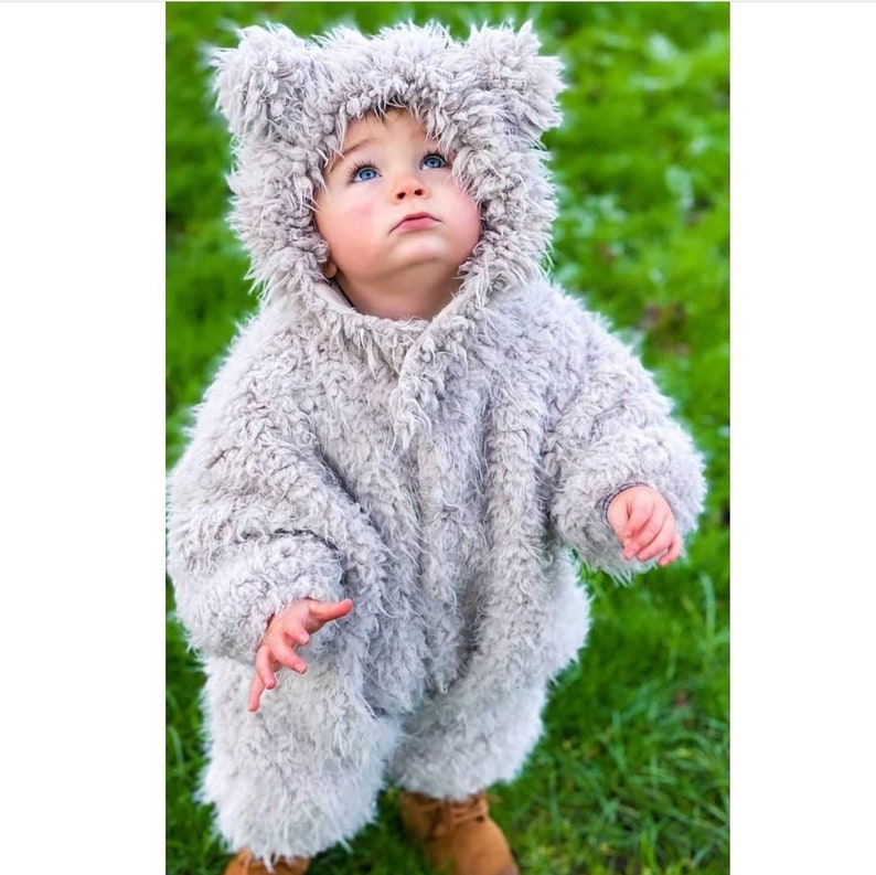 May include: A baby wearing a gray, furry bear costume with a hood and brown boots stands on green grass. The costume has a button closure and bear ears on the hood. The baby is looking up.