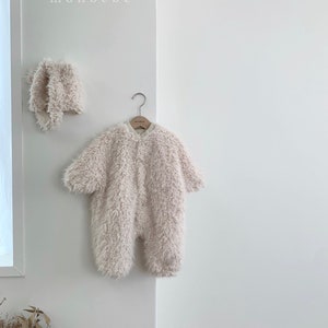 May include: A fluffy, light beige baby jumpsuit and matching bonnet hang on a white wall. The jumpsuit has long sleeves and a button-up front. The word "monbebe" is printed in small, white letters above the items.