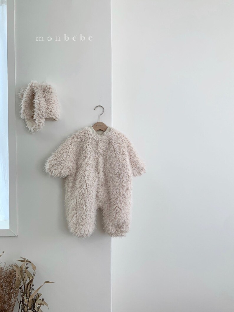 May include: A fluffy, light beige baby jumpsuit and matching bonnet hang on a white wall. The jumpsuit has long sleeves and a button-up front. The word "monbebe" is printed in small, white letters above the items.