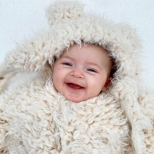 May include: A smiling baby wearing a cream-colored, fluffy hooded outfit. The outfit has a button closure and a soft, textured appearance. The baby's face is visible, with a wide smile and rosy cheeks, creating a warm and inviting image.