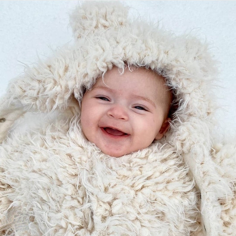May include: A smiling baby wearing a cream-colored, fluffy hooded outfit. The outfit has a button closure and a soft, textured appearance. The baby's face is visible, with a wide smile and rosy cheeks, creating a warm and inviting image.