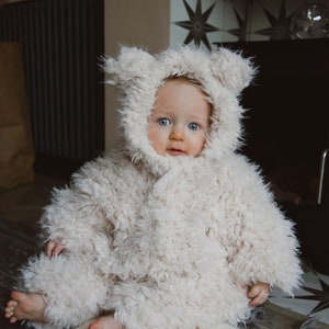 May include: A baby wearing a fluffy, cream-colored hooded coat with bear ears. The coat is made of a soft, textured material. The baby has blue eyes and is sitting on a light-colored rug. The background includes a dark cabinet and patterned tile.