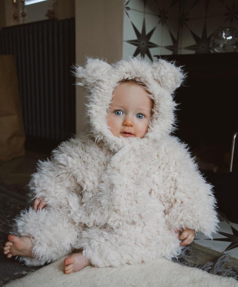 May include: A baby wearing a fluffy, cream-colored hooded coat with bear ears. The coat is made of a soft, textured material. The baby has blue eyes and is sitting on a light-colored rug. The background includes a dark cabinet and patterned tile.