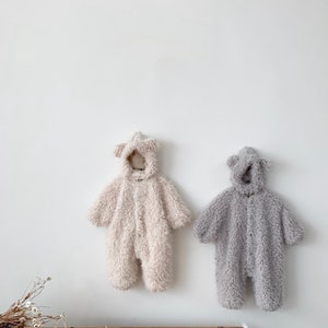 May include: Two plush baby jumpsuits hanging on a white wall. One is a light beige color, and the other is gray. Both have attached hoods with bear ears and button closures down the front. The jumpsuits are made of a soft, fuzzy material.