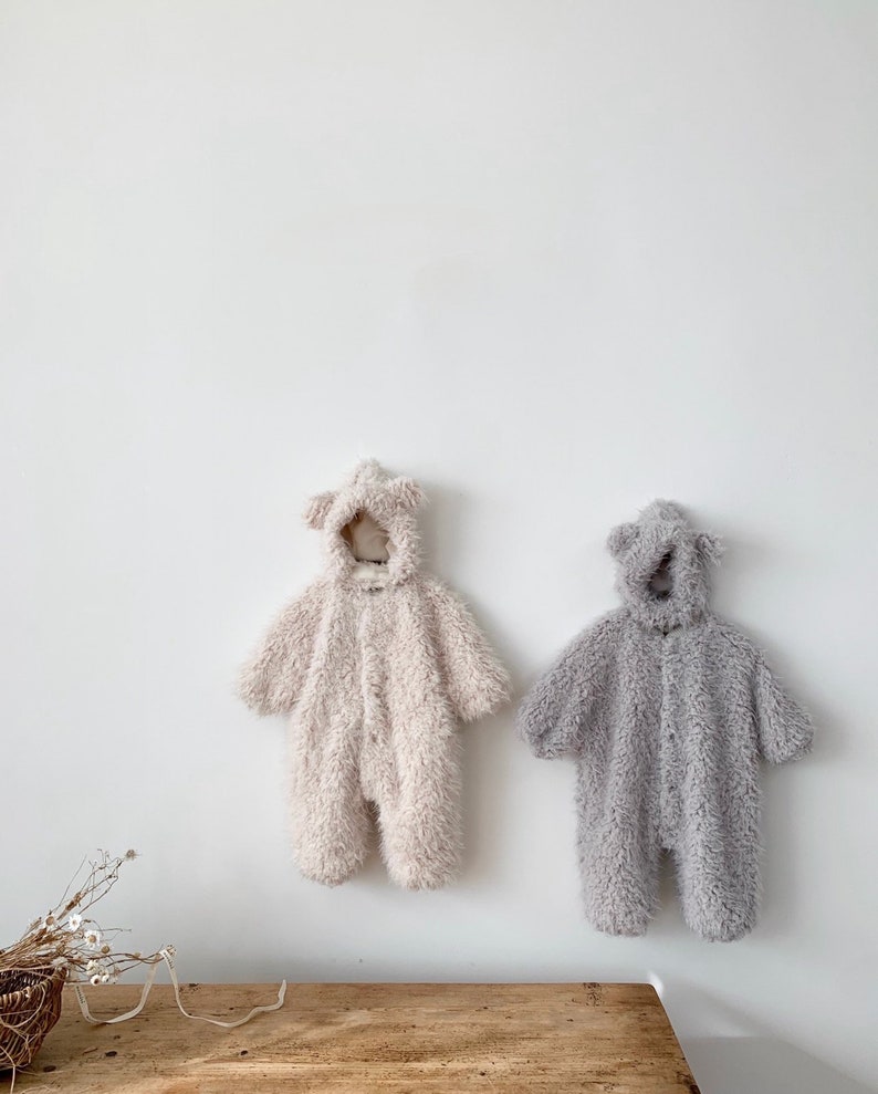 May include: Two plush baby jumpsuits hanging on a white wall. One is a light beige color, and the other is gray. Both have attached hoods with bear ears and button closures down the front. The jumpsuits are made of a soft, fuzzy material.