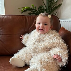 May include: A smiling baby wearing a fluffy, cream-colored jumpsuit with matching socks, sitting on a brown leather couch. The baby has a small bow in their hair. A houseplant is visible in the background.