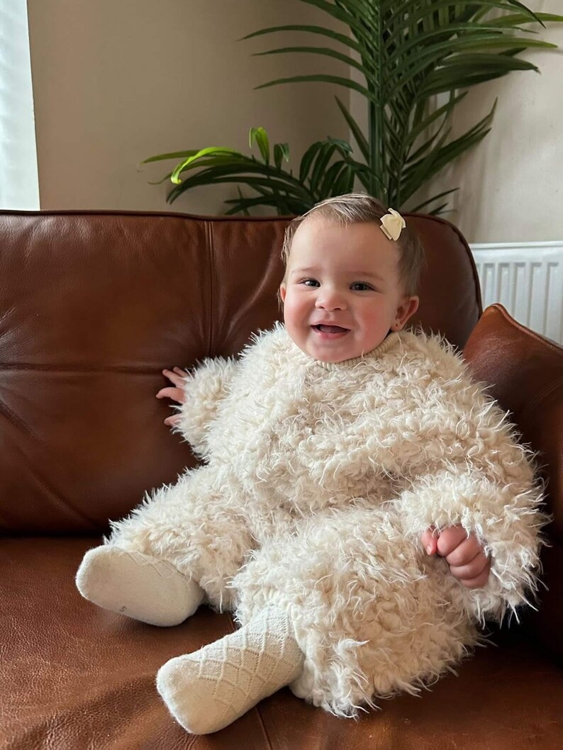 May include: A smiling baby wearing a fluffy, cream-colored jumpsuit with matching socks, sitting on a brown leather couch. The baby has a small bow in their hair. A houseplant is visible in the background.