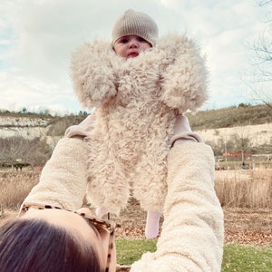 May include: A baby wearing a beige, fluffy, teddy bear-style jumpsuit and a matching knit cap is held up against a cloudy sky. The jumpsuit has a hood and covers the baby's arms and legs. The person holding the baby is wearing a cream-colored jacket.