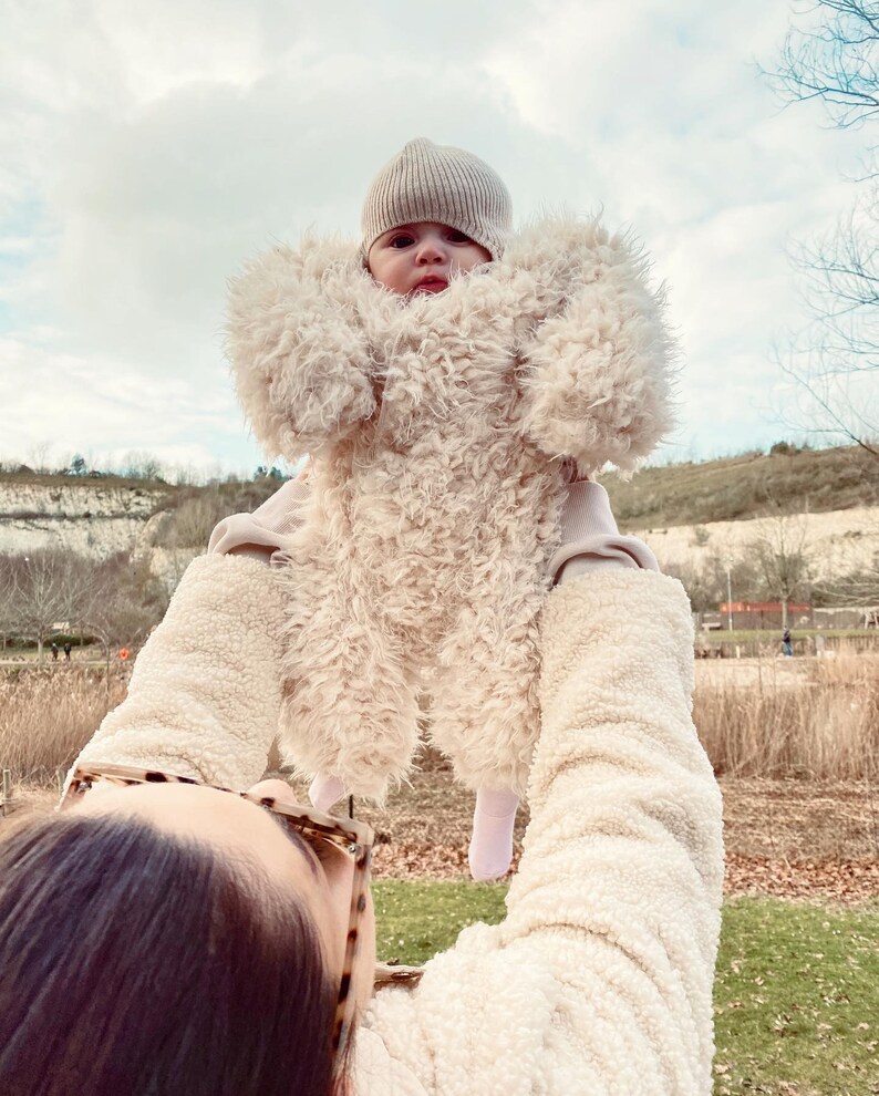 May include: A baby wearing a beige, fluffy, teddy bear-style jumpsuit and a matching knit cap is held up against a cloudy sky. The jumpsuit has a hood and covers the baby's arms and legs. The person holding the baby is wearing a cream-colored jacket.