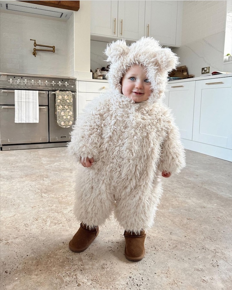 May include: A child wearing a cream-colored bear costume with a hood and matching furry jumpsuit. The costume has bear ears and is paired with brown boots. The child is standing in a kitchen with white cabinets and a stainless steel oven.