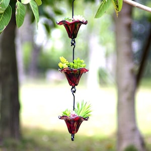 May include: A hanging, three-tiered planter with red and black glass flower-shaped pots. Each pot holds small green and yellow plants. The planter hangs from a tree branch with a black metal chain.