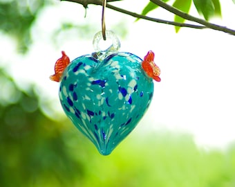 Pode incluir: Um bebedouro para beija-flores de vidro azul em forma de coração com detalhes vermelhos e brancos pendurado em um galho.