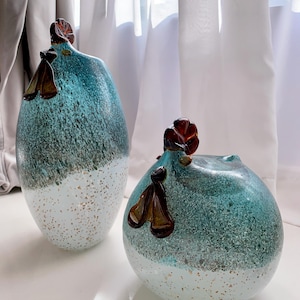 May include: Two decorative glass vases. The vases are teal and white with a speckled pattern. Each vase has a brown glass accent on top. The vases are different sizes and shapes, one is oval and the other is round.