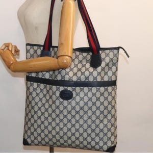 May include: A vintage tote bag with a repeating pattern in beige and navy blue. The bag features navy blue leather trim and handles with red and navy stripes. A navy blue emblem is centered on the front of the bag.