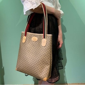 May include: A brown and beige tote bag with a brown leather trim and a red, green, and white striped strap. The bag has a GG pattern and a Gucci logo.