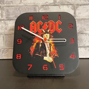 Könnte beinhalten: Schwarze quadratische Uhr mit einem rot-weißen AC/DC-Logo und einem Bild von Angus Young. Das Zifferblatt der Uhr hat rote Zahlen und weiße Zeiger.