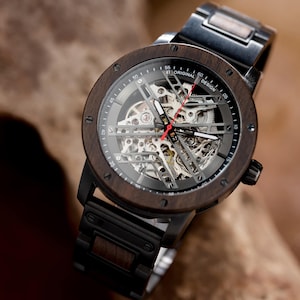 May include: A brown wooden wristwatch with a black metal band and a skeletonized clock face. The watch face has a silver background with black markings and a red second hand. The watch face also has the text "ORIGINAL DESIGN" printed on it.