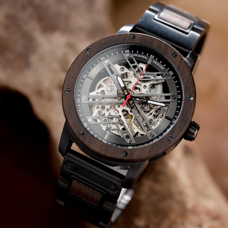 May include: A brown wooden wristwatch with a black metal band and a skeletonized clock face. The watch face has a silver background with black markings and a red second hand. The watch face also has the text "ORIGINAL DESIGN" printed on it.