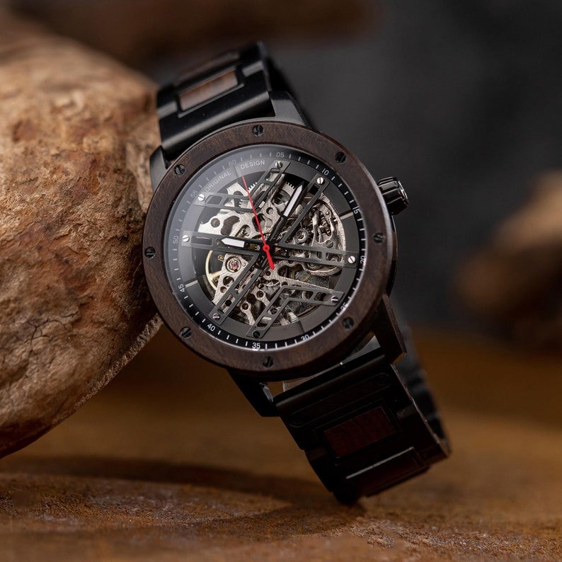 May include: A black and brown wooden wristwatch with a skeletonized automatic movement. The watch face has white markings and the text "ORIGINAL DESIGN" printed around the edge.