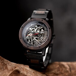 May include: A black wooden wristwatch with a black metal band and a skeletonized automatic movement. The watch face has white markings and the text "ORIGINAL DESIGN" printed on it.