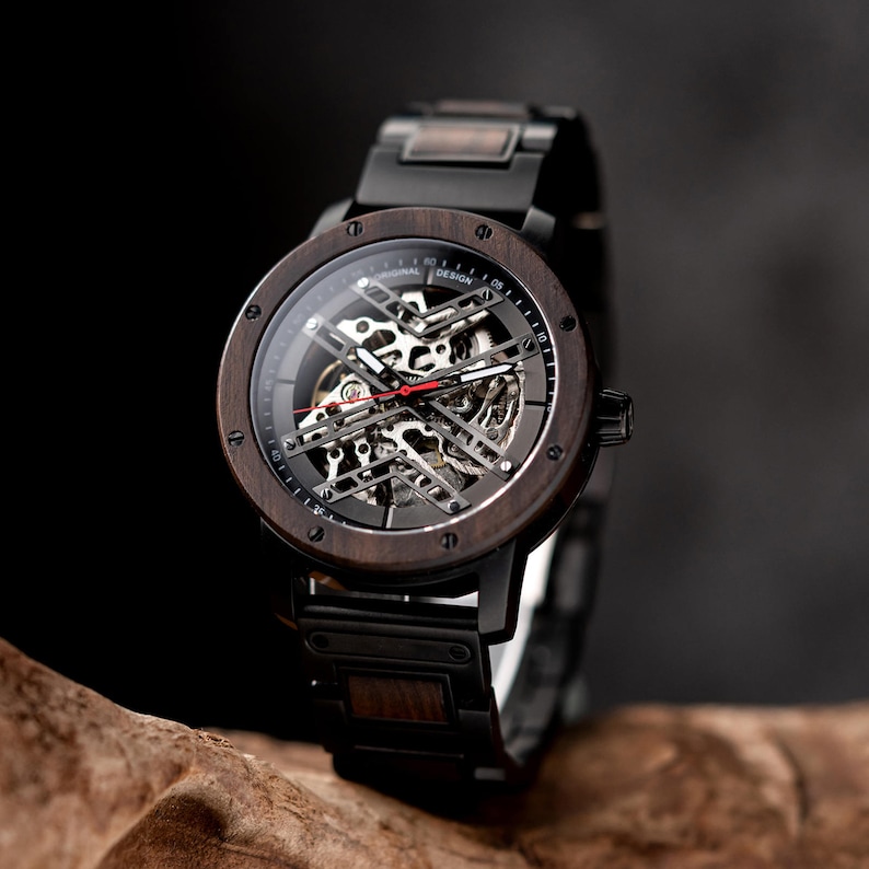 May include: A black wooden wristwatch with a black metal band and a skeletonized automatic movement. The watch face has white markings and the text "ORIGINAL DESIGN" printed on it.