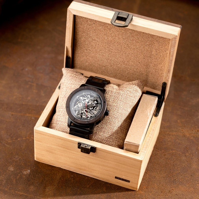 May include: A black wooden watch with a skeletonized movement is displayed in a wooden box with a hinged lid. The box is made of light wood with a natural finish.