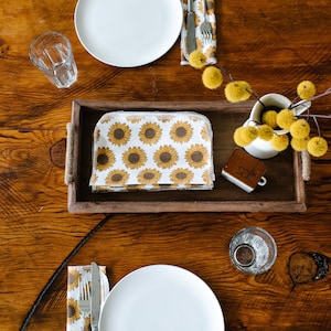 May include: A wooden table set for two with white plates, silverware, and glasses.  The table is decorated with a sunflower patterned napkin, a small wooden sign that says "Salt", and a vase with yellow flowers.