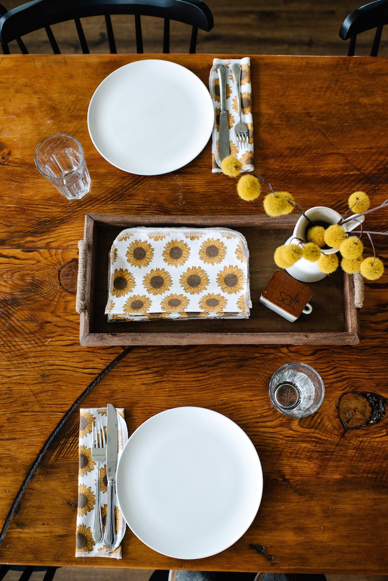 May include: A wooden table set for two with white plates, silverware, and glasses.  The table is decorated with a sunflower patterned napkin, a small wooden sign that says "Salt", and a vase with yellow flowers.