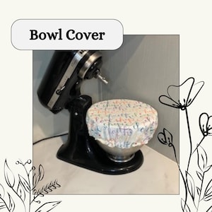 以下が含まれることがあります： 黒いスタンドミキサーとボウルカバー。ボウルカバーは白で、カラフルな花柄と「love, hope, faith」の文字がプリントされています。「Bowl Cover」の文字は白いボックスに入っています。