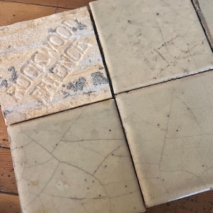 Può includere: Primo piano di quattro piastrelle quadrate bianco sporco dall'aspetto usurato. Una piastrella presenta il testo in rilievo "ROCKWOOD TRENTON". Le piastrelle mostrano una rete di sottili crepe e macchie scure.