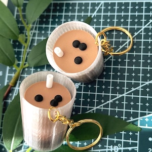 May include: Two miniature bubble tea keychains. Each keychain features a cup with a brown liquid, black boba pearls, and a white straw. The keychains have gold-colored rings and are set against a green cutting mat.