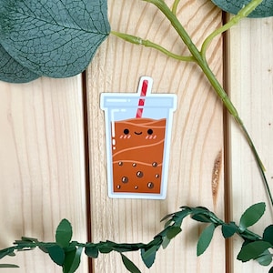 May include: A brown and white cartoon illustration of a cup of boba tea with a red and white straw. The boba tea has a smiling face and is surrounded by brown boba pearls.