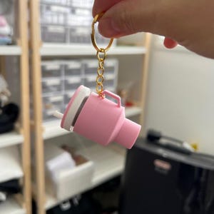 Może przedstawiać: Różowy miniaturowy brelok do kubka z białą pokrywką i słomką. Brelok jest przymocowany do złotego pierścienia i łańcuszka. Kubek ma mały uchwyt. Tło jest rozmyte, pokazując półki i czarne urządzenie.