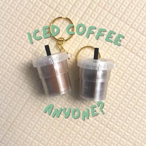 May include: Two miniature iced coffee keychains with gold key rings. One keychain has a brown coffee color, the other is gray. The text "ICED COFFEE ANYONE?" is in green above and below the keychains.