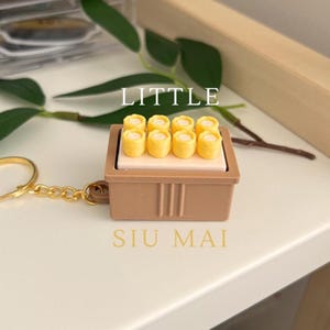 Könnte beinhalten: Ein Miniatur-Siu-Mai-Schlüsselanhänger. Der Schlüsselanhänger zeigt einen braunen rechteckigen Behälter mit acht gelben Siu-Mai-Knödeln obenauf. Der Text "LITTLE" steht über dem Behälter und "SIU MAI" darunter. Eine goldfarbene Kette ist befestigt.