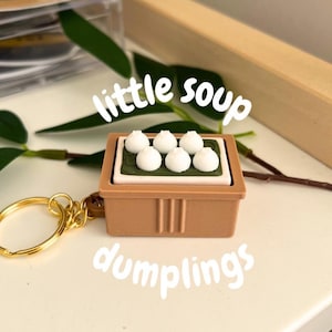 Peut inclure: Un porte-clés en forme de raviolis bruns avec un anneau doré. Le porte-clés représente une petite boîte brune avec six raviolis blancs sur un tapis vert. Les mots "little soup dumplings" sont écrits en blanc.